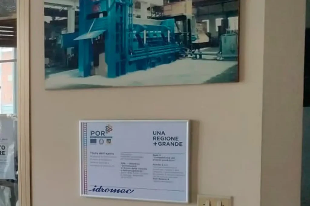 Targhetta POR FESR esposta all’ingresso dello stabilimento Idromec spa, con foto storica della pressa